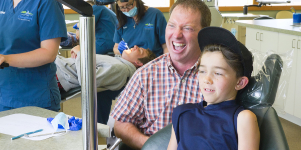 There is a father and son sitting at the dental clinic and smiling behind are the doctors who are attending to patients